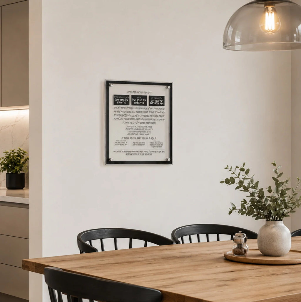 A framed Hebrew and English prayer plaque with a black border hangs on a white wall in a modern dining room setting with a wooden table, black chairs, and a hanging glass dome lamp.