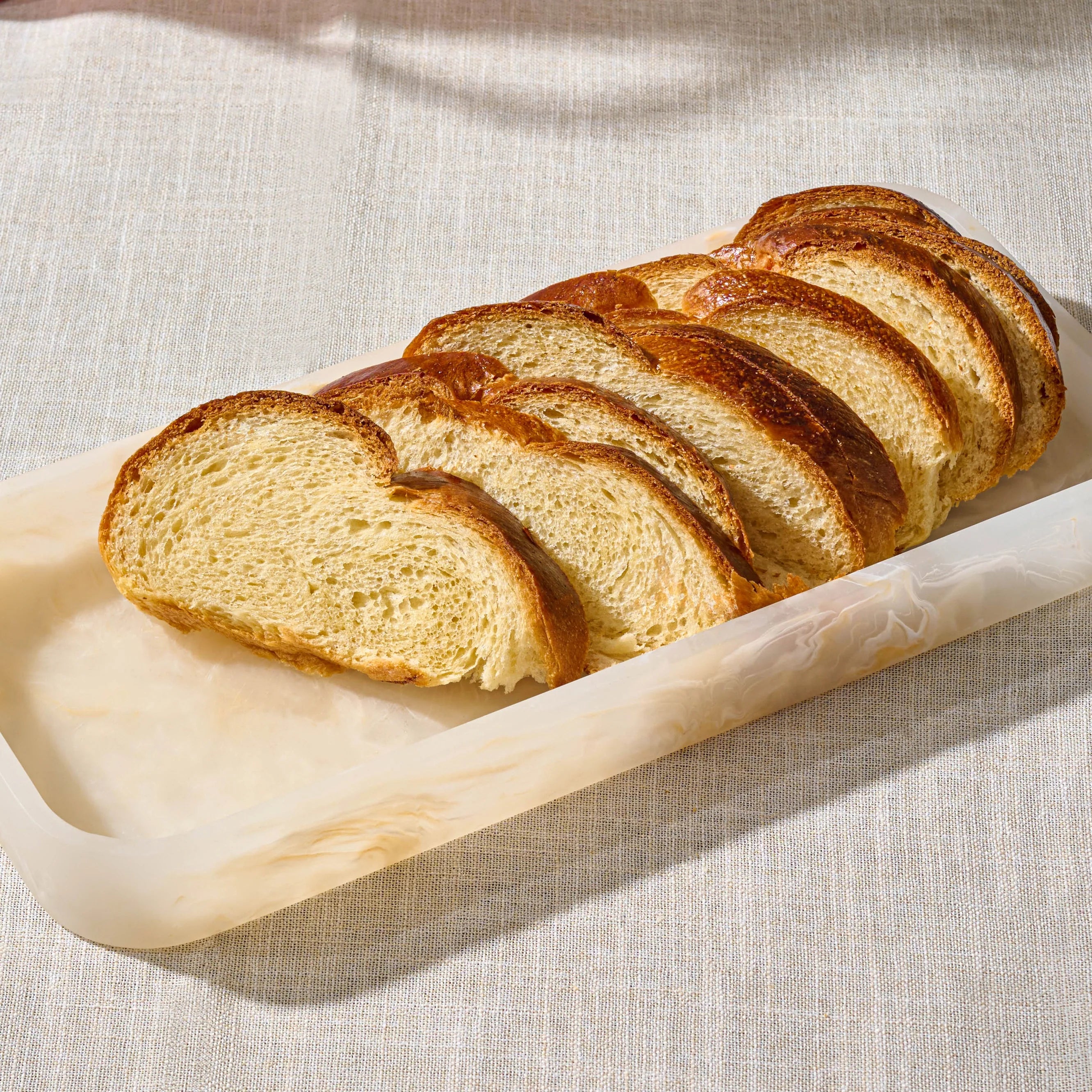 Slices of golden-brown artisan bread arranged diagonally on a translucent rectangular servingresin tray over a textured beige fabric surface.