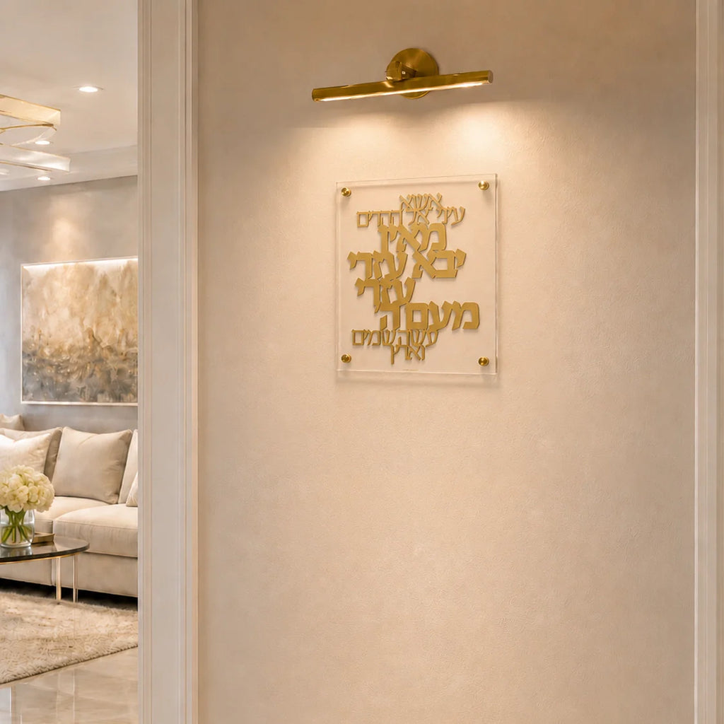 Gold Hebrew letter wall art mounted on a beige textured wall, illuminated by a gold wall sconce. The acrylic plaque is shown in a modern, elegant living room setting.