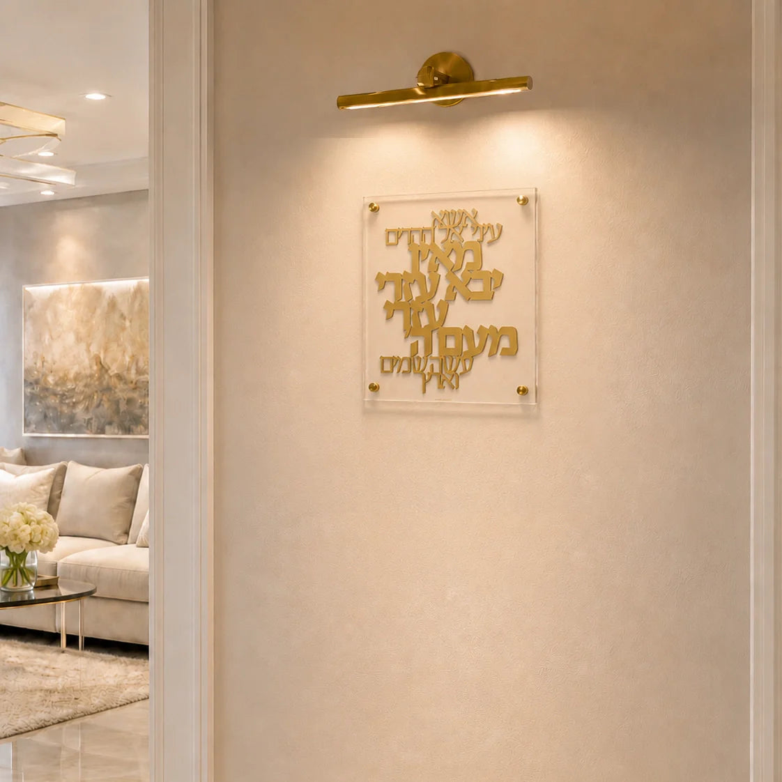 Gold Hebrew letter wall art mounted on a beige textured wall, illuminated by a gold wall sconce. The acrylic plaque is shown in a modern, elegant living room setting.