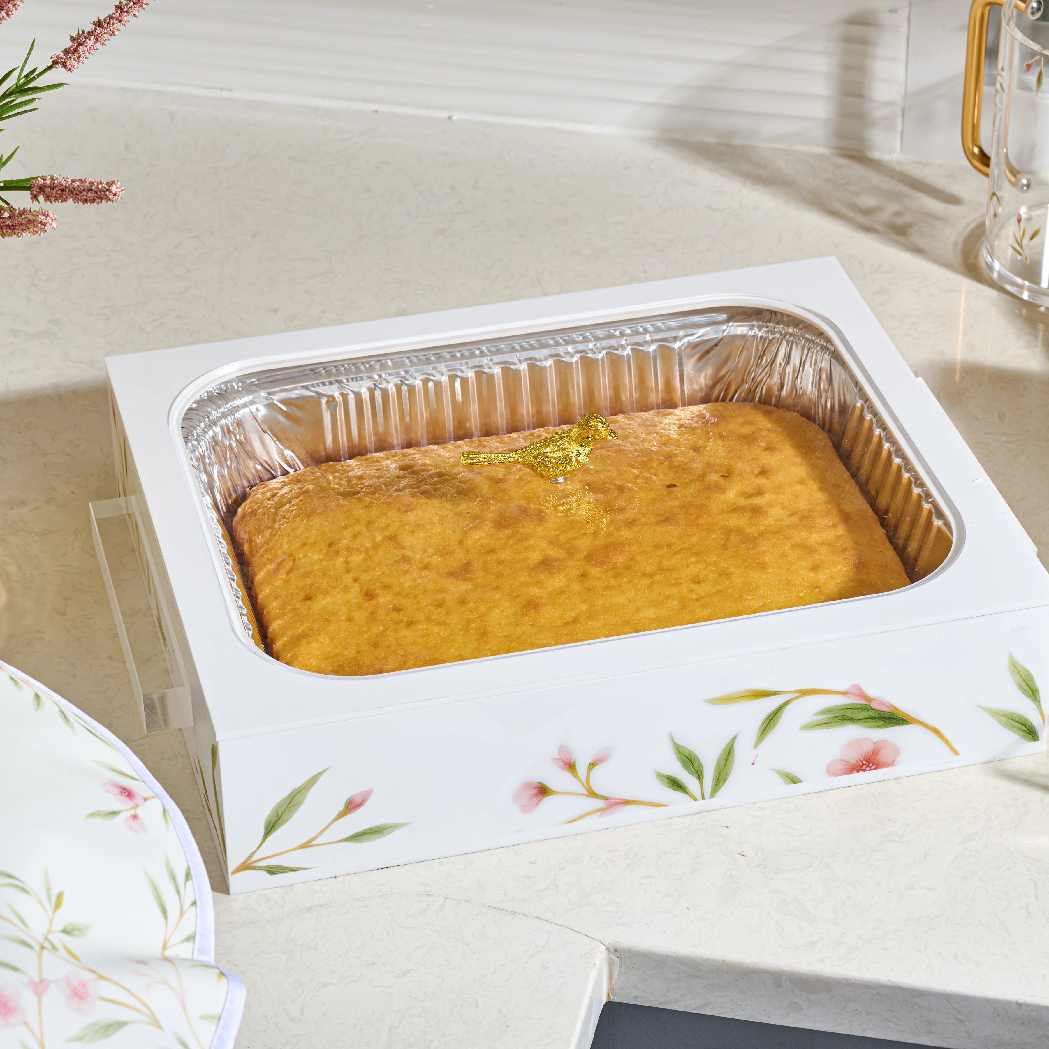 A rectangular golden-brown baked cake or casserole presented in a white floral acrylic tray with a silver foil-lined bottom, set on a light countertop.