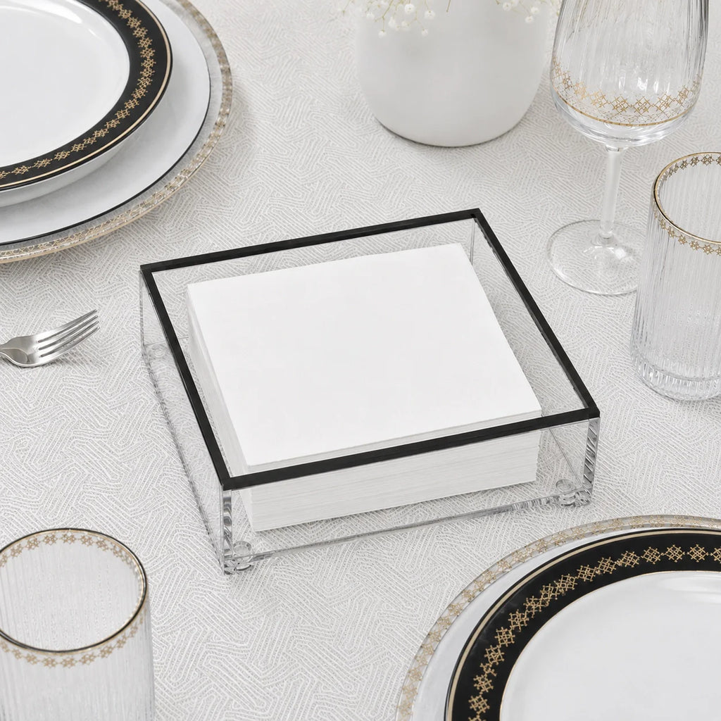 A clear square napkin holder with a black frame, filled with white folded napkins, styled on a white tablecloth with gold-rimmed plates, glasses, and a fork.