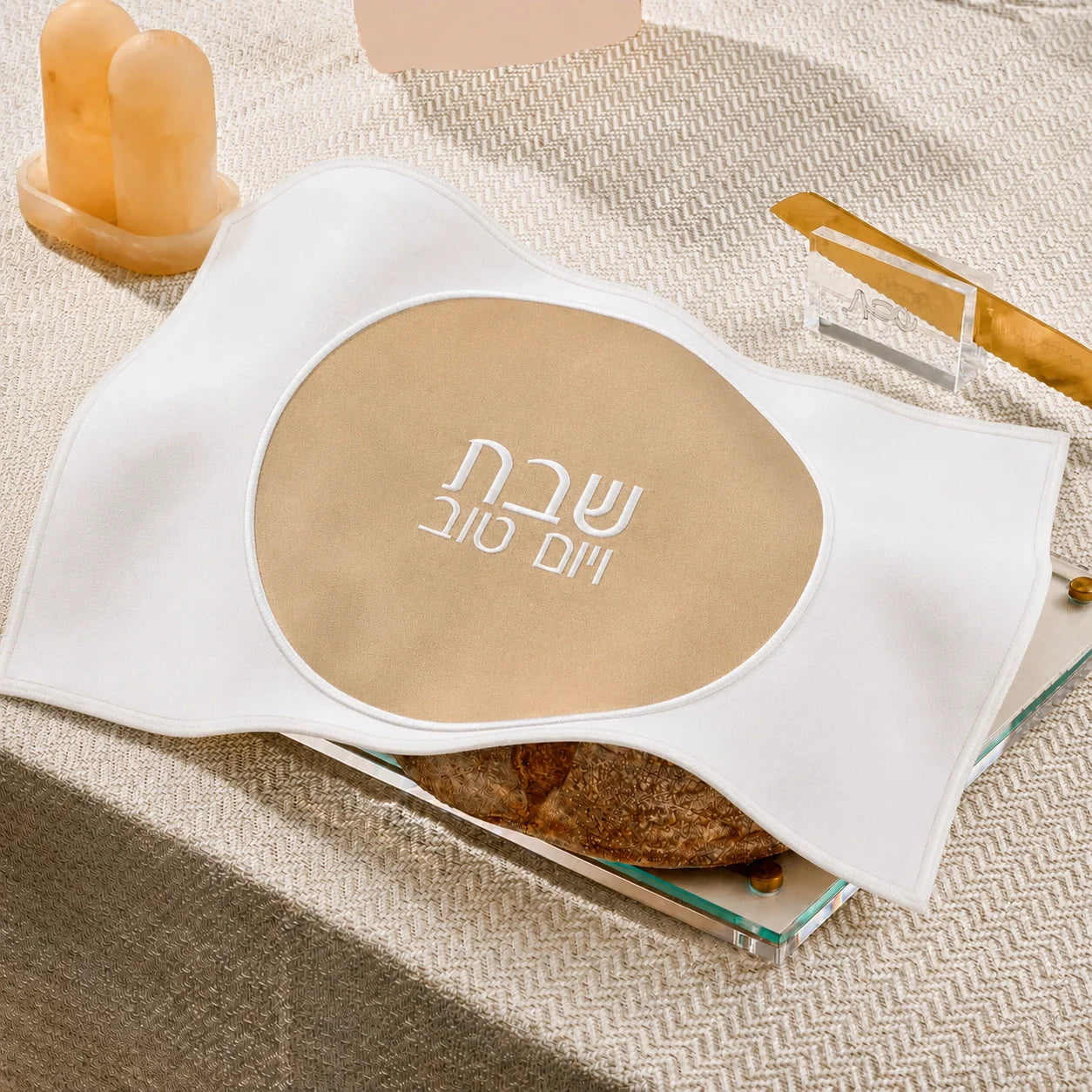 A white fabricchallah  cover with a large beige oval center printed with white Hebrew  text, styled on a textured beige surface with candles and a small dish in the background.