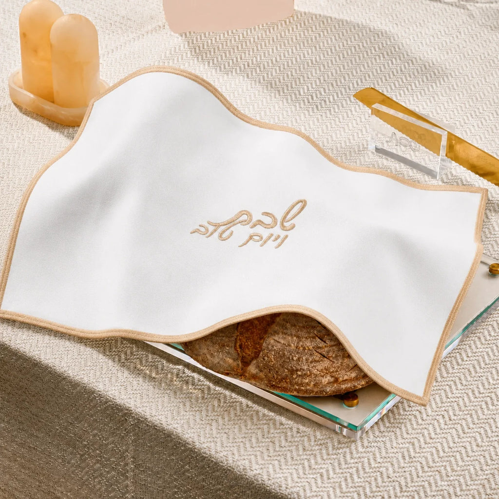 A white fabric napkin with a beige border and Arabic calligraphy embroidery, draped over a stack of books with a loaf of bread visible underneath, on a textured beige tablecloth.