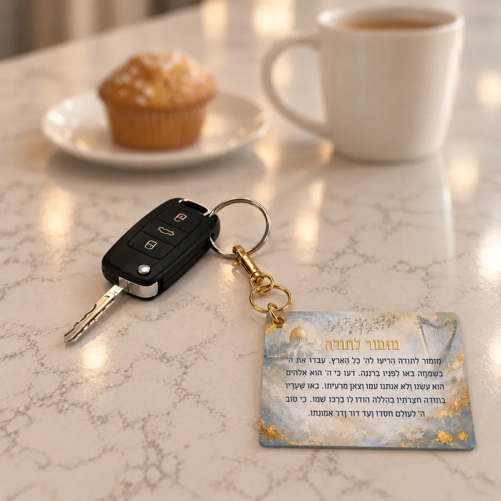 A painted leatherette Mizmor Lesoda keychain with a harp, musical notes and a Jerusalem dome in gold and grey, on a gold clasp. On a marble kitchen counter. 
