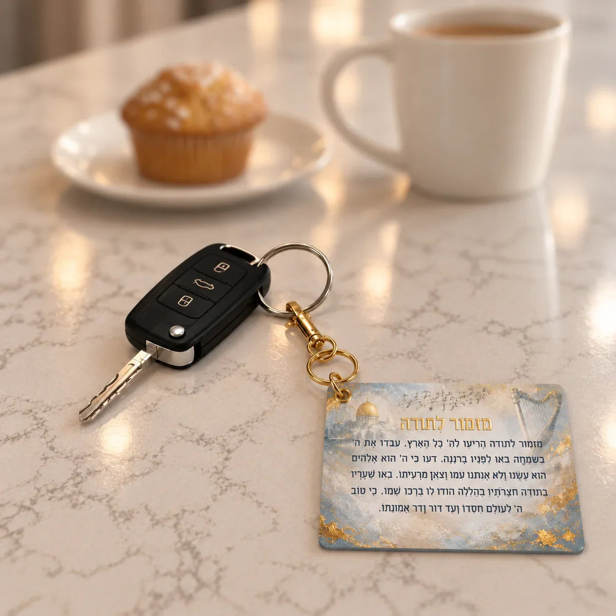 A painted leatherette Mizmor Lesoda keychain with a harp, musical notes and a Jerusalem dome in gold and grey, on a gold clasp. On a marble kitchen counter. 