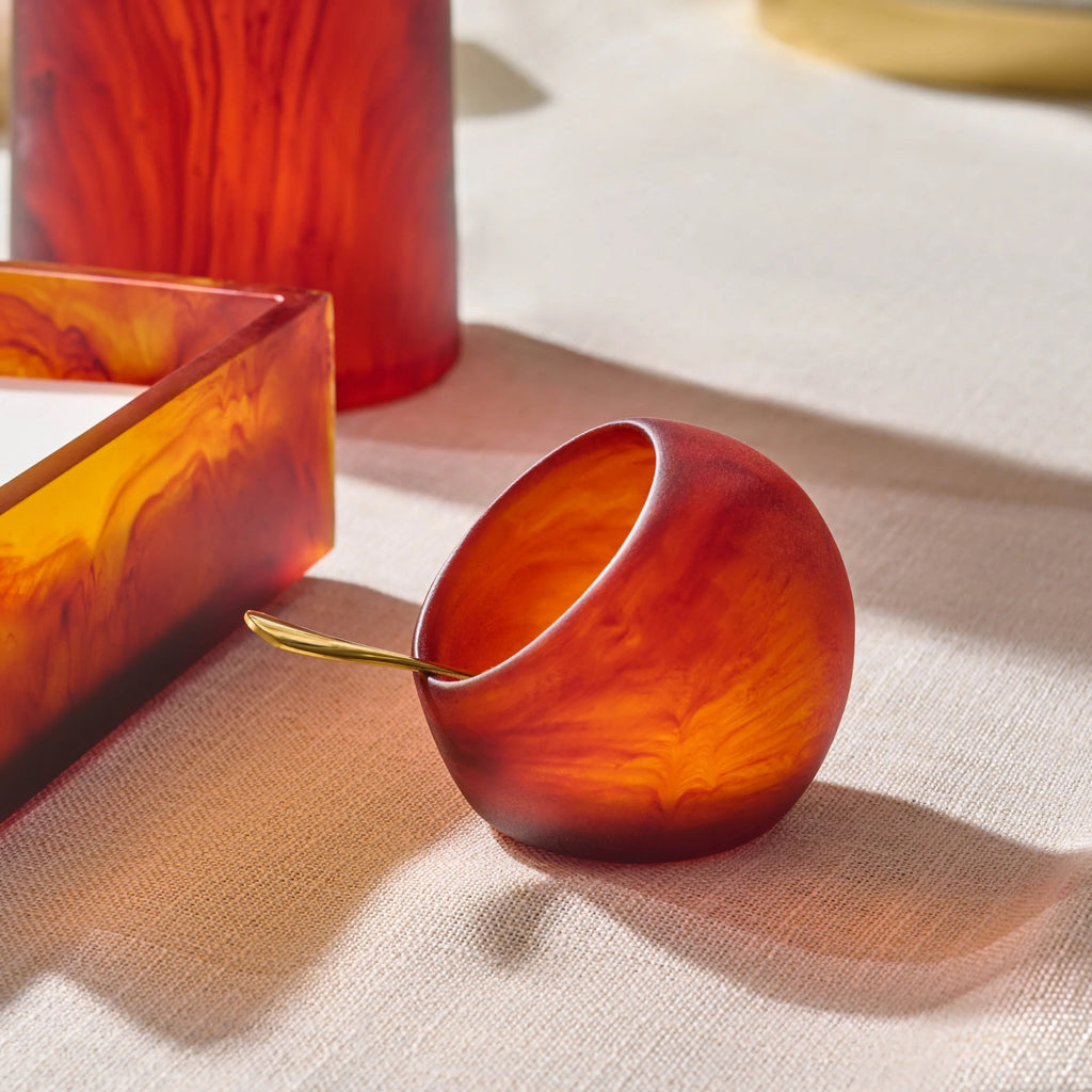 A small rounded amber-orange salt cellar  with a gold spoon resting inside, styled on a beige fabric surface with matching amber glassware in the background.