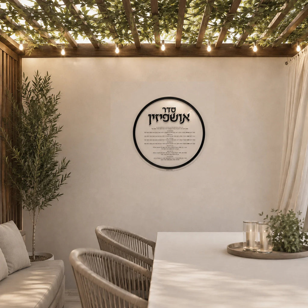 Round clear Lucite Ushpizin sukkah decoration hanging in a modern succah with a Sukkot table, chairs, and potted plants.