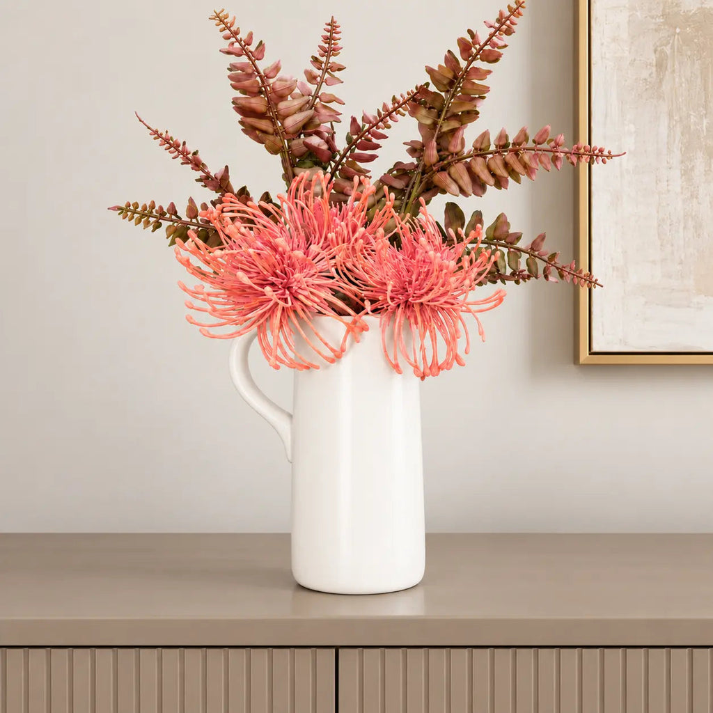 White ceramic pitcher vase with coral faux flowers styled on a fluted console beside a gold framed canvas