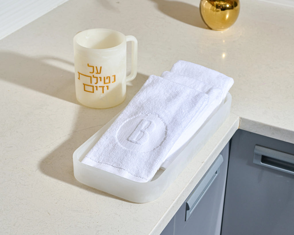 A white hand towel with an embroidered monogram 'B' inside a circle, folded in a translucent white resin holder on a light beige countertop next to a cream-colored resin mug with gold Hebrew lettering.