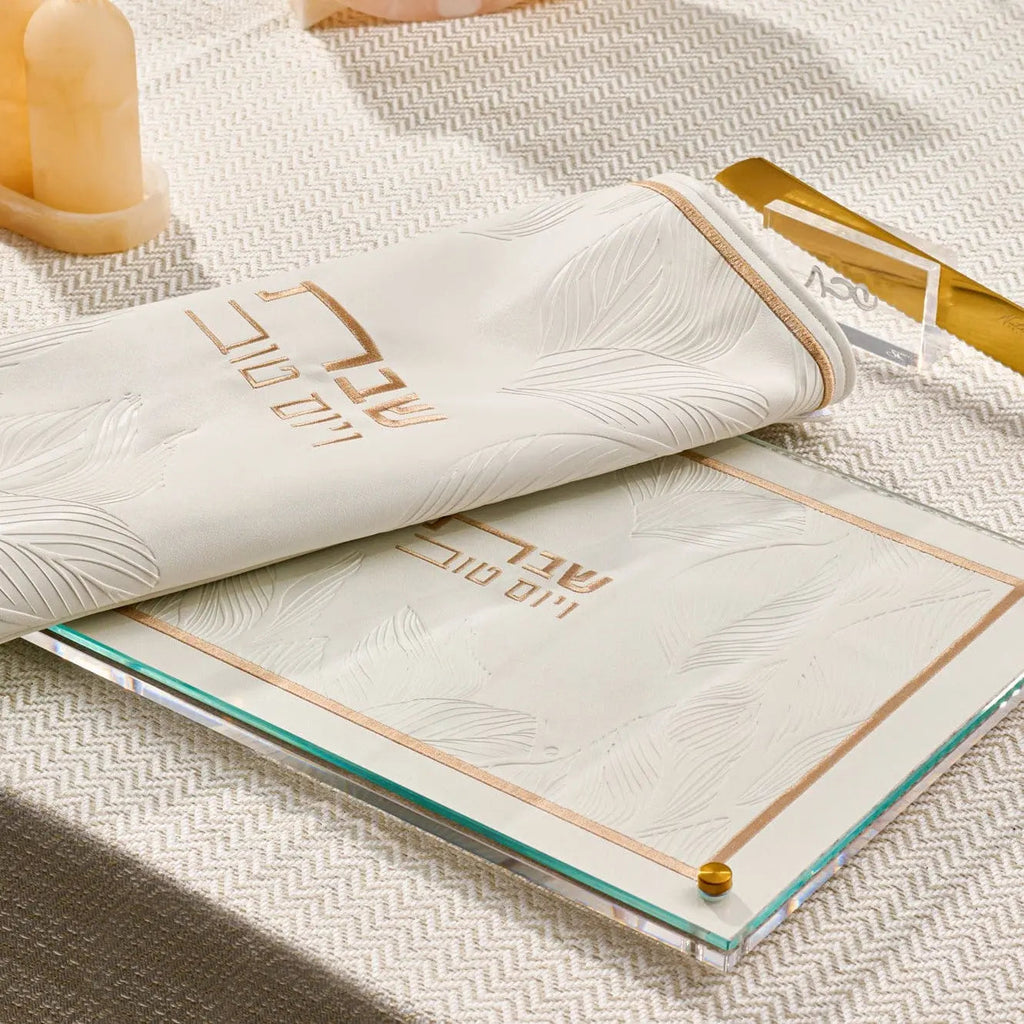 Embossed leaf challah board in gold and black leather by Waterdale Collection, displayed with a white napkin and gold utensils on a textured surface.