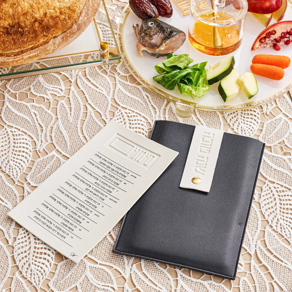 Black leather Simanim pouch with lucite cards displayed on white tablecloth, styled with holiday table setting and refreshments.