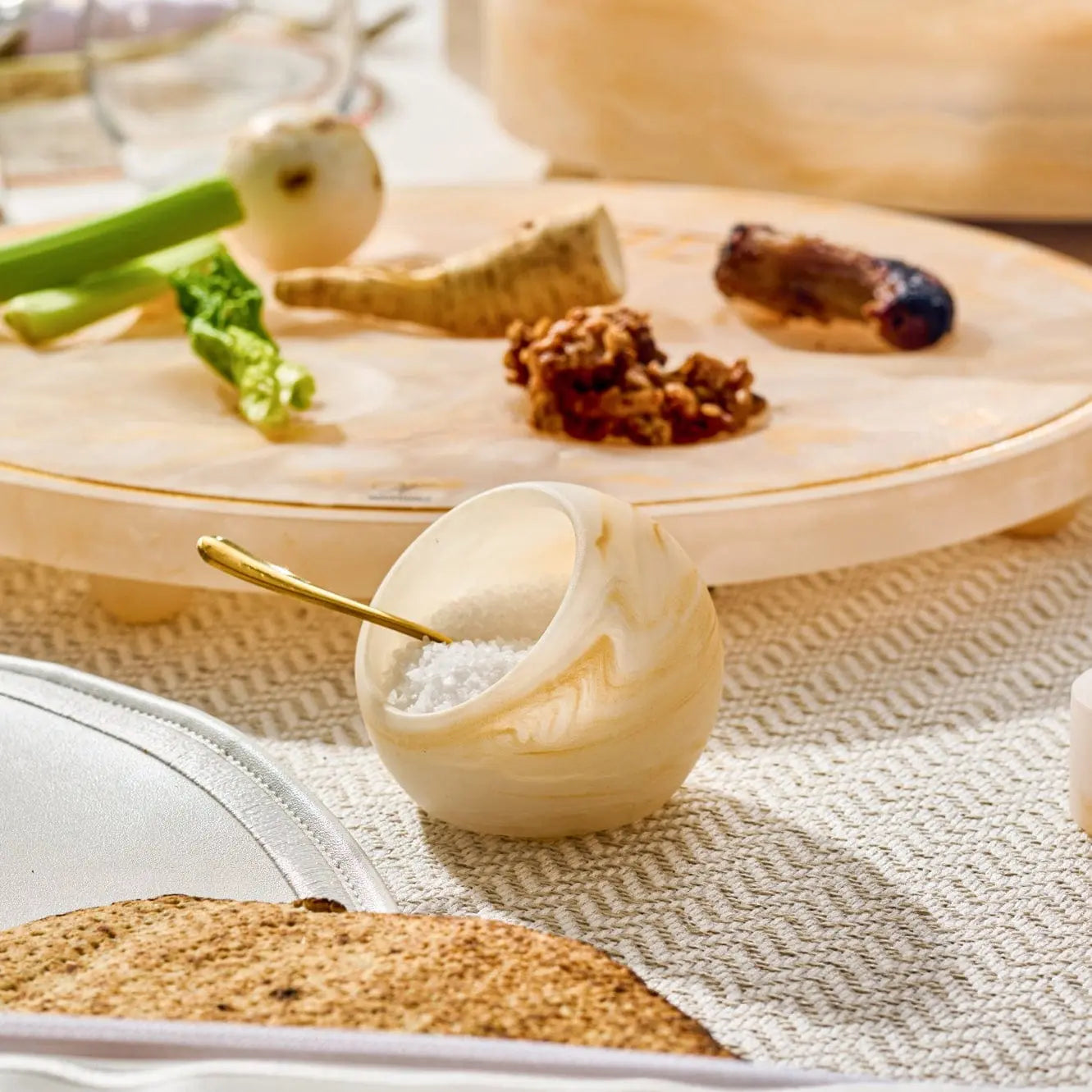 Cream resin salt cellar with gold spoon sitting on neutral linens, surrounded by fresh herbs and spices on wooden board.