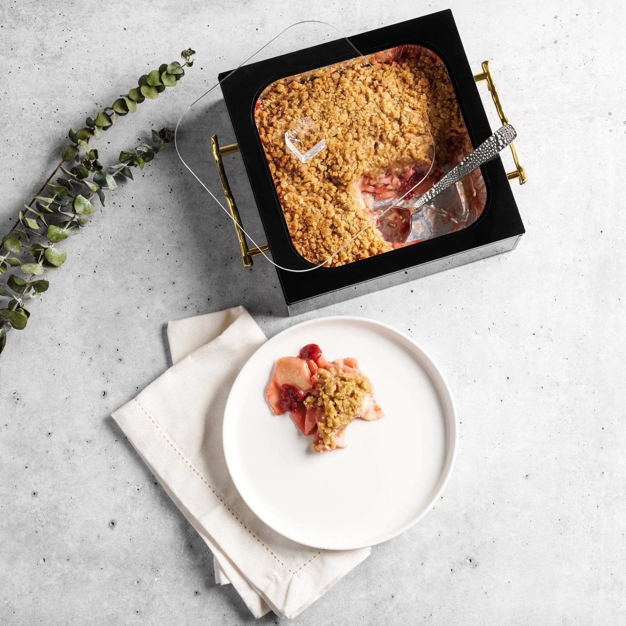 Black square 8x8 pan holder displaying baked good with fresh berries, styled with eucalyptus and white plate on marble surface.