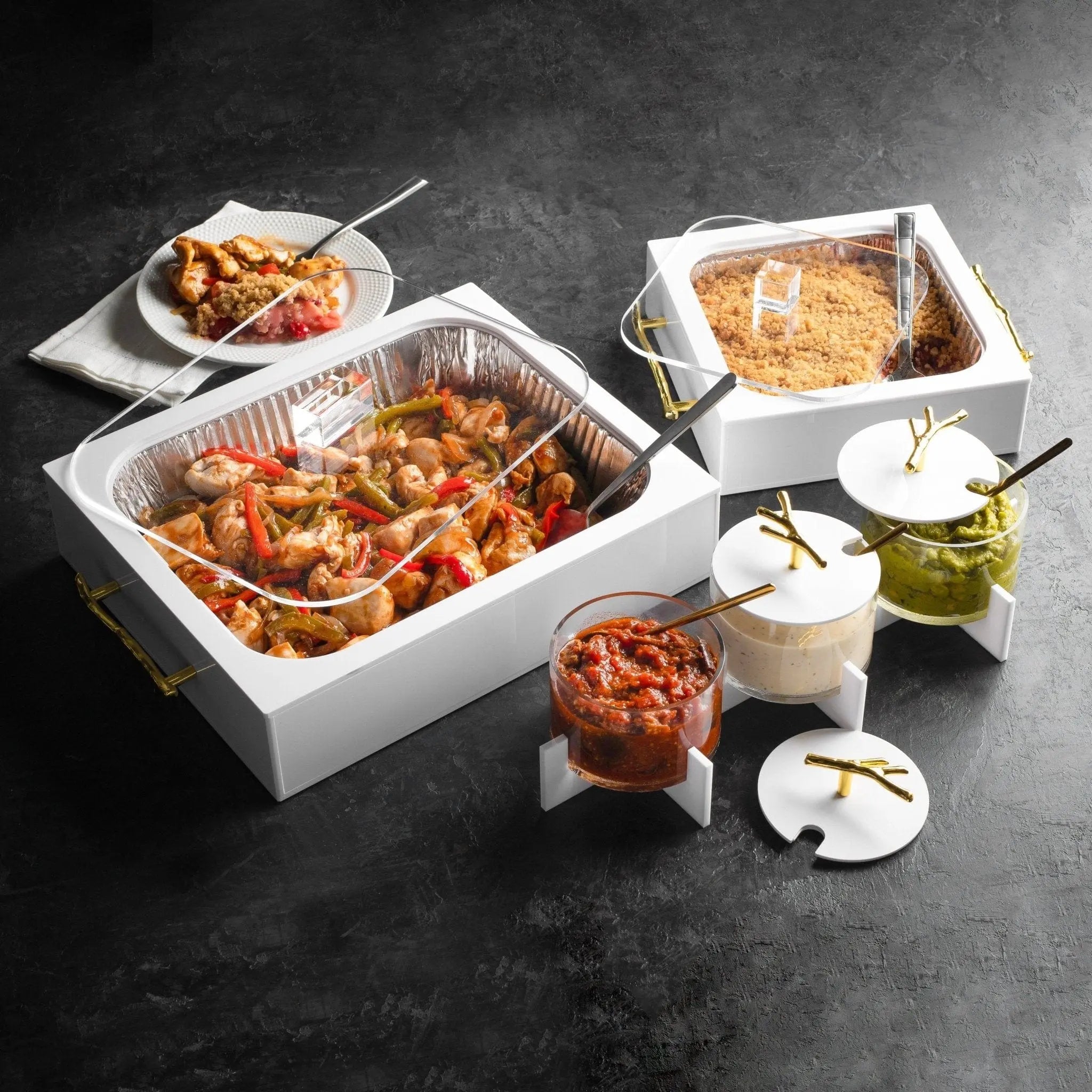 White ceramic 8x8 pan holder displaying appetizers and baked goods on dark surface with serving utensils