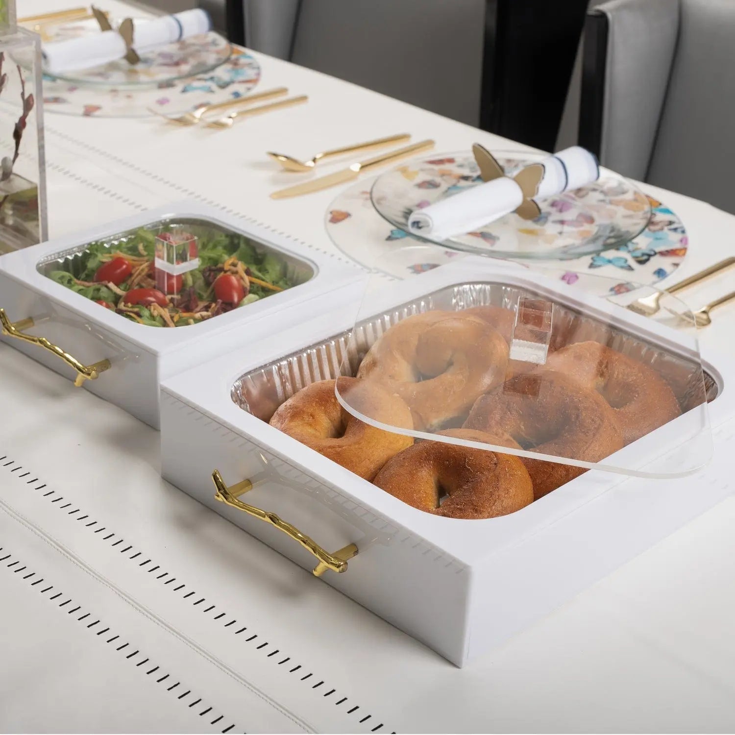 White 8x8 pan holder with gold trim displaying prepared food in clear disposable containers on a styled tabletop.