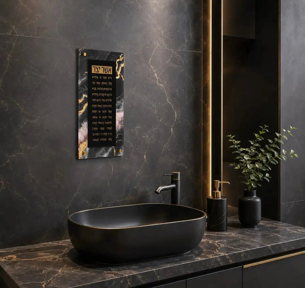 Black agate floating wall art displaying Asher Yatzar text, mounted above a modern bathroom sink with gold accents and marble countertop.
