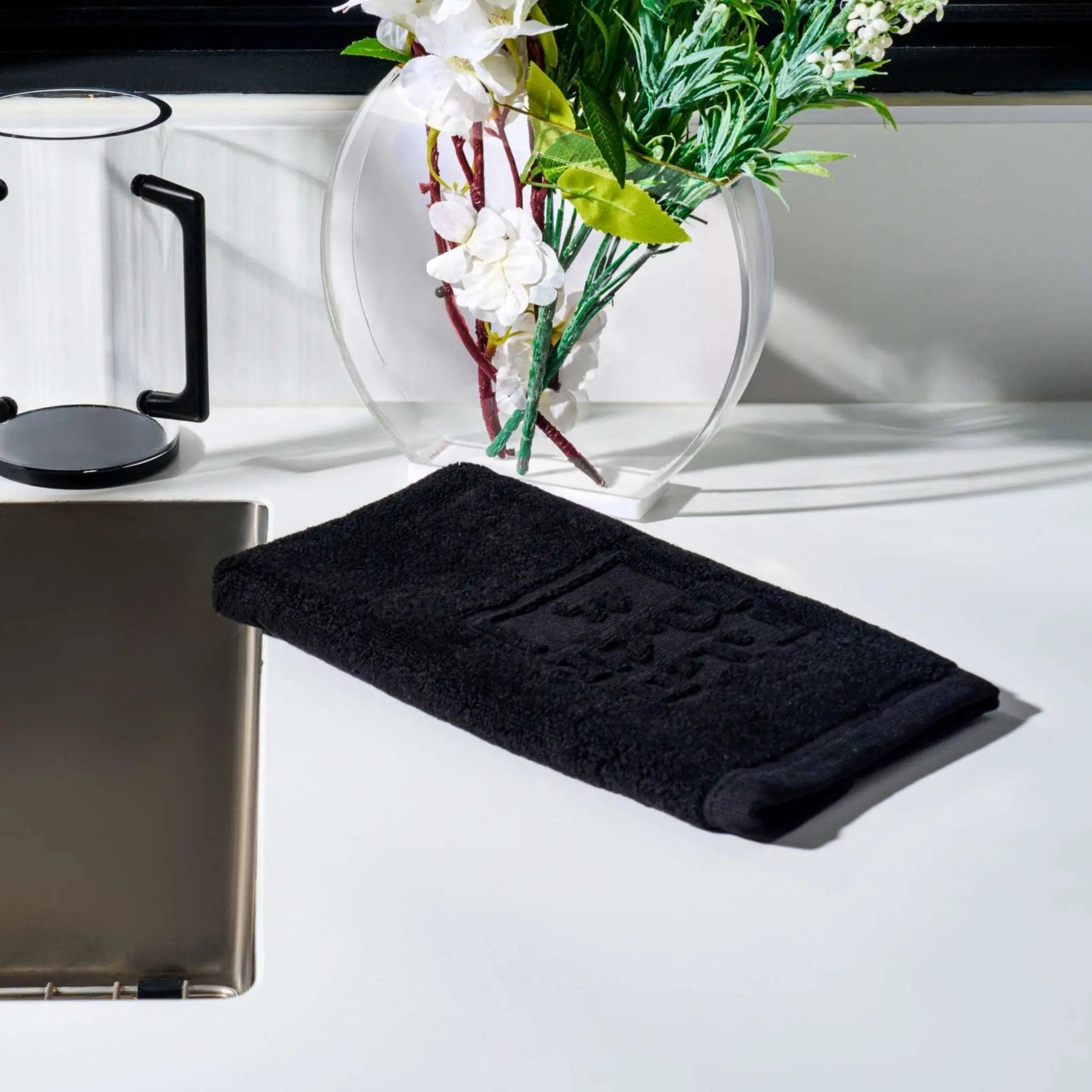 Black embossed finger towel for netilas yadayim displayed on white surface next to flowers and coffee mug.