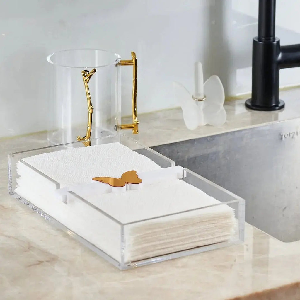 White marble butterfly paper towel holder with brass accents displayed on a bathroom vanity counter next to toiletries and a sink.