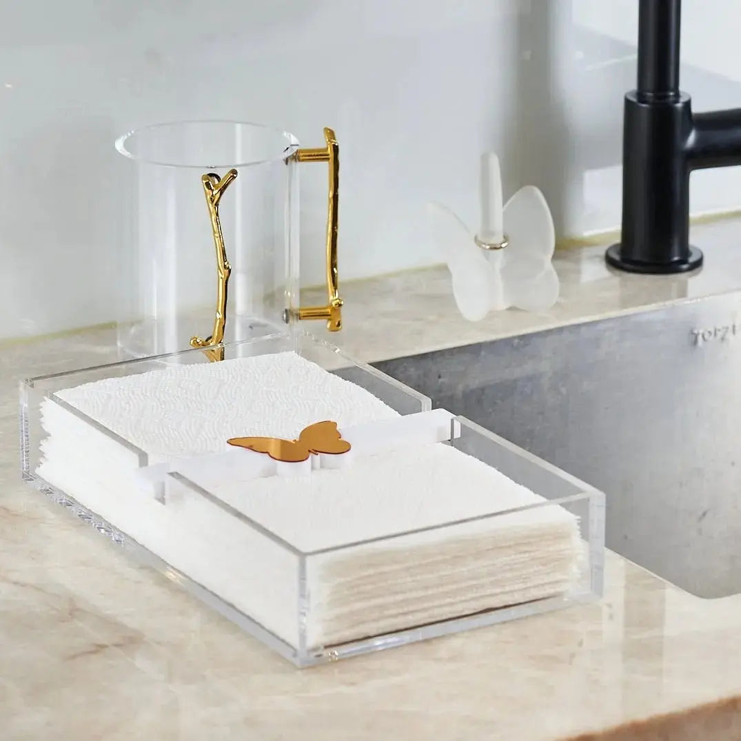 White marble butterfly paper towel holder with brass accents displayed on a bathroom vanity counter next to toiletries and a sink.