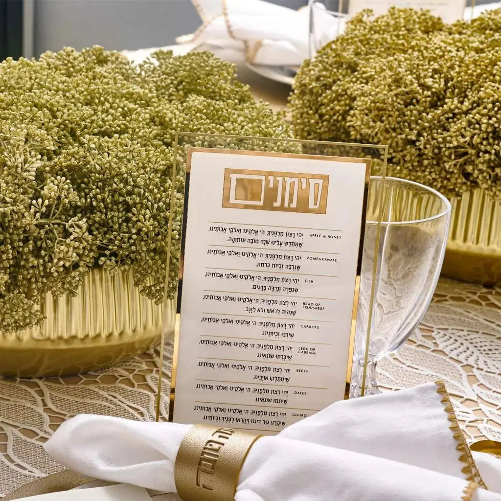 Classic 2.0 Simanim Card in clear lucite with gold accents, displayed with white napkin and boxwood landscaping backdrop
