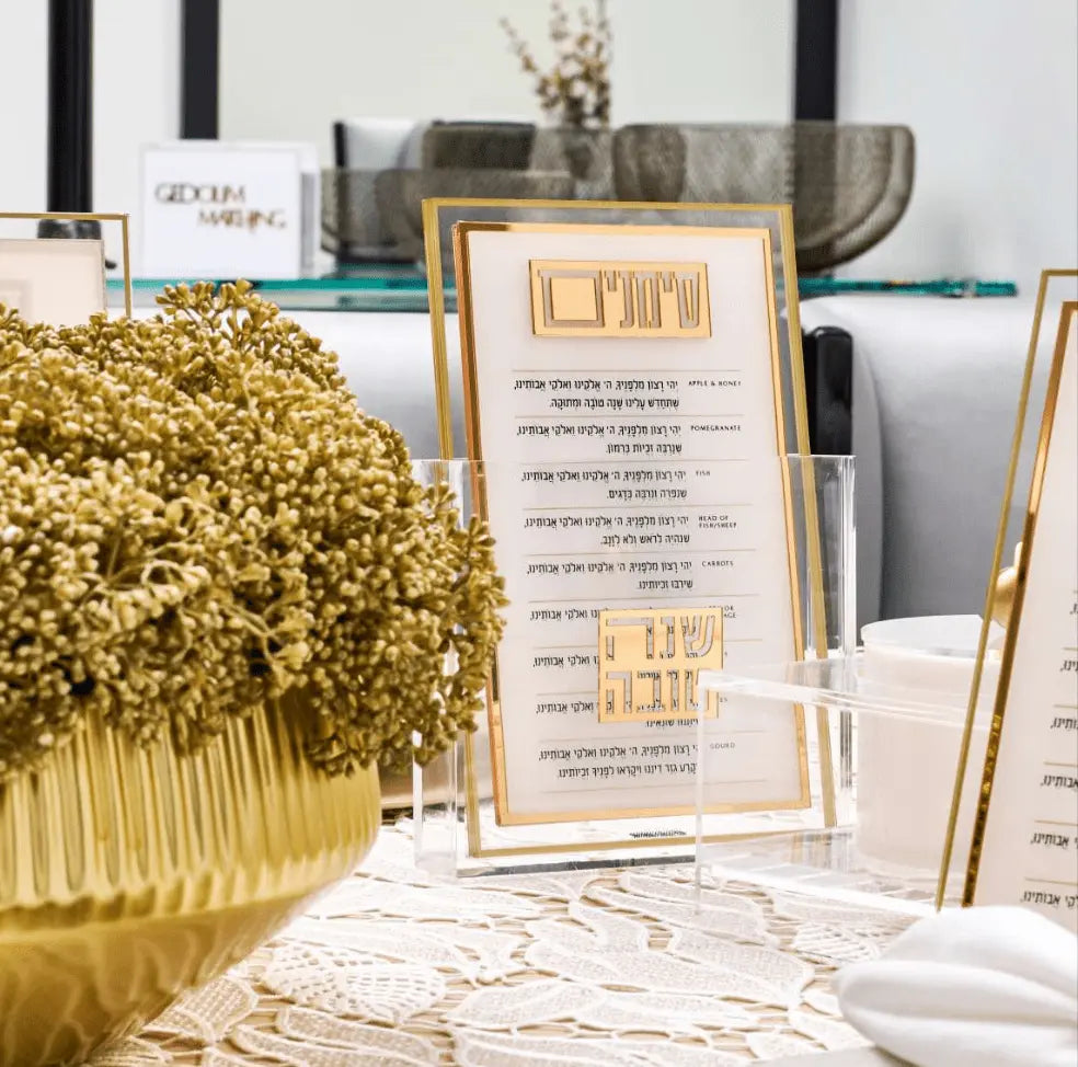 Classic 2.0 Simanim Card Set in clear lucite with gold text, displayed with gold floral arrangement and white linens