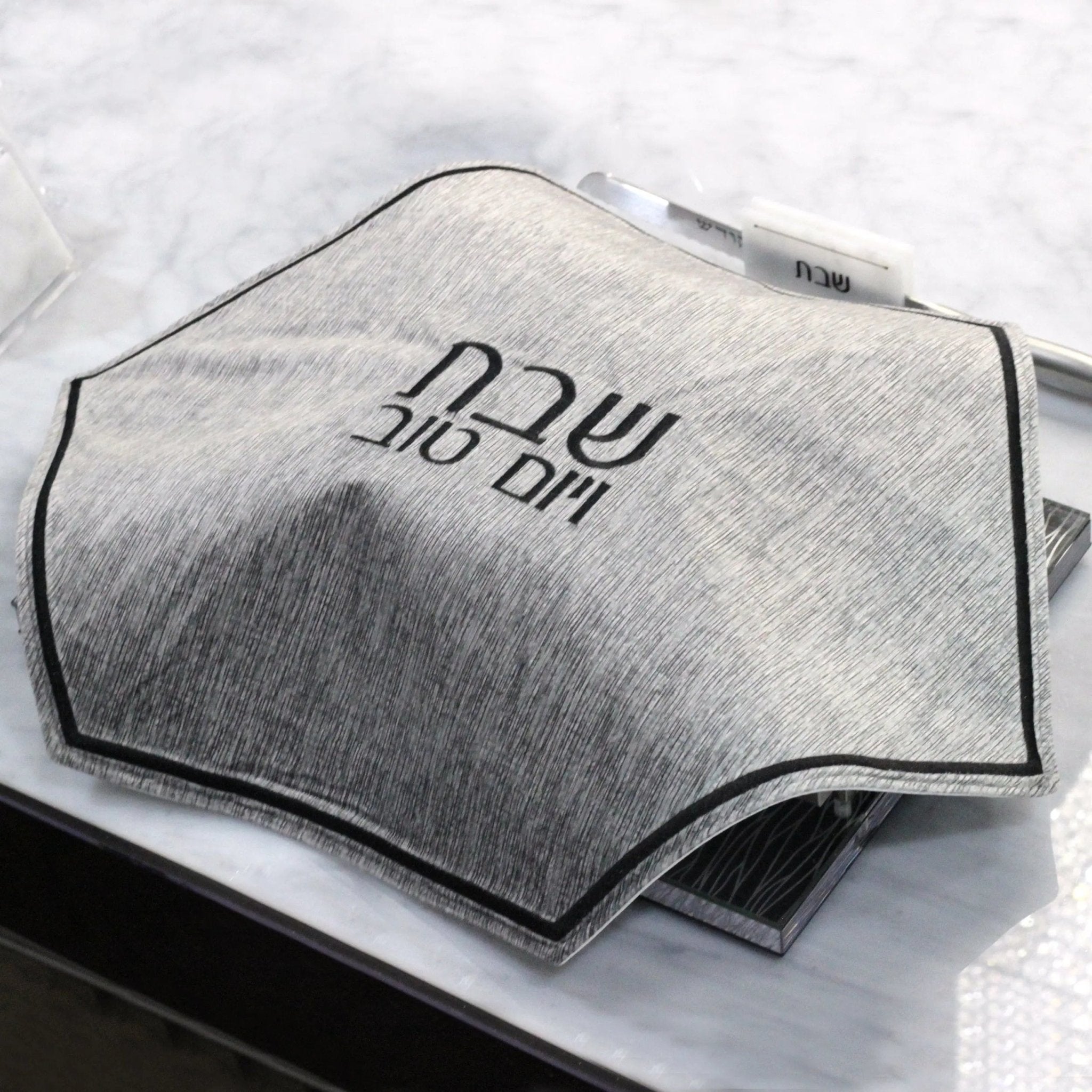 Gray crushed velvet hexagon challah cover with embroidered Hebrew text, displayed on white fabric background.