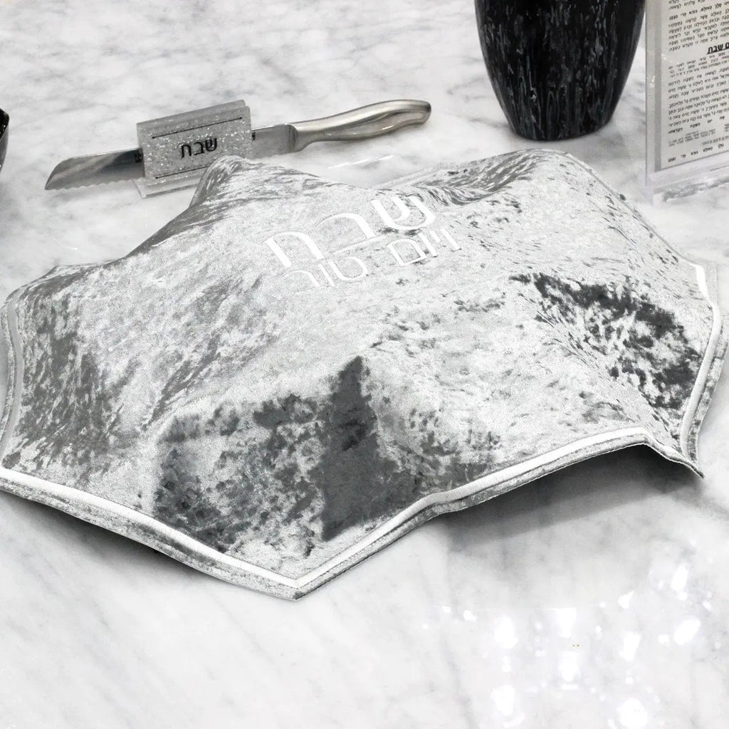 Crushed velvet hexagon challah cover in black with silver accents displayed on white marble surface with challah knife nearby.