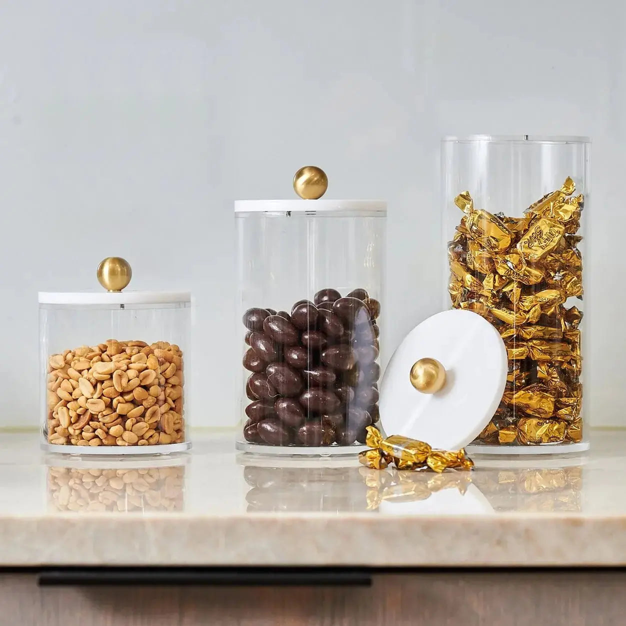 Four clear cylinder canisters with brass lids holding cereal, chocolate chips, pasta, and cornflakes on a marble surface.
