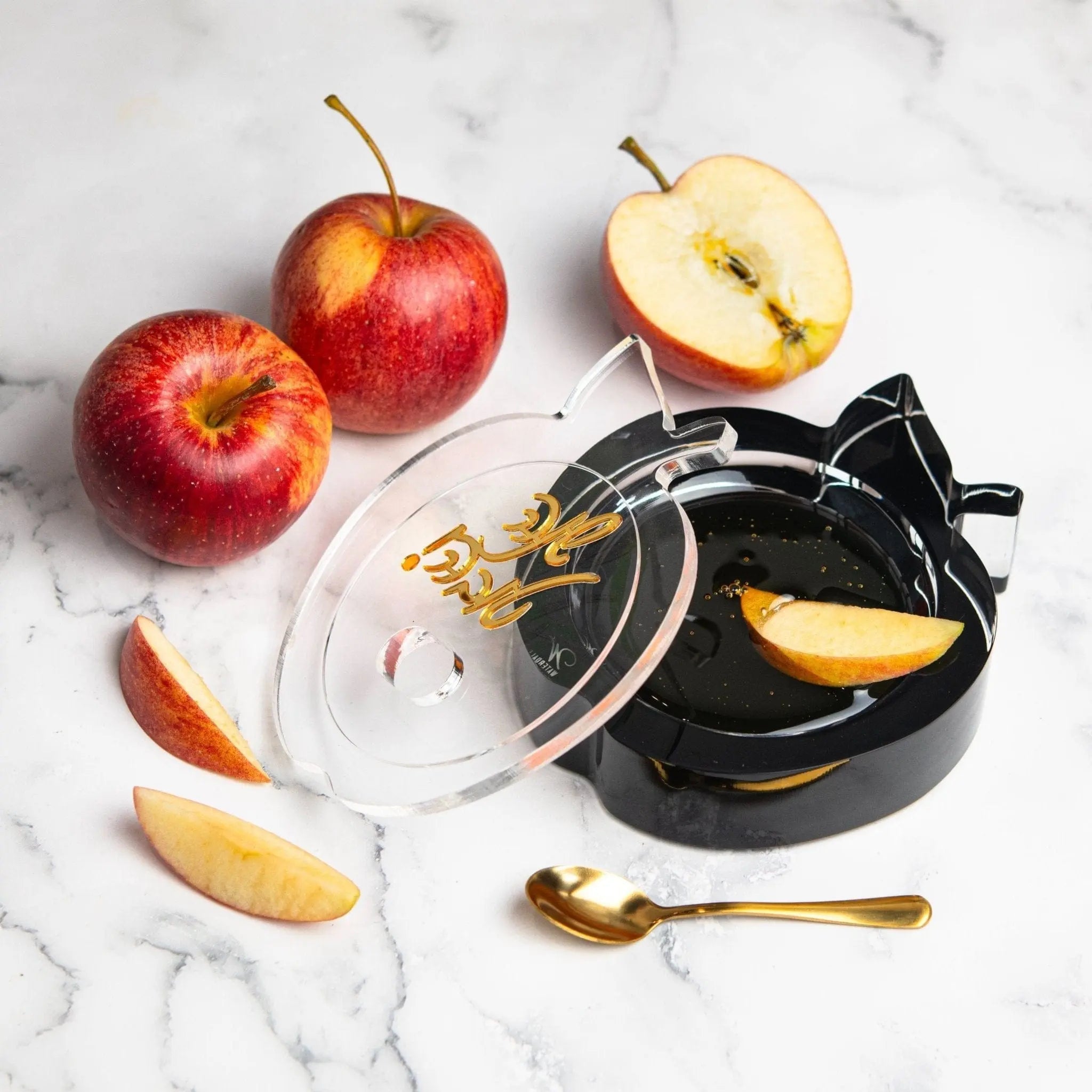 Red apple-shaped honey dish with black handle beside fresh apples and wooden honey dipper on marble surface.