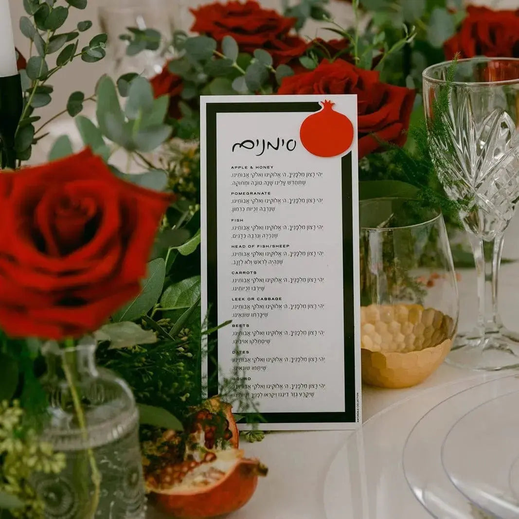 Red pomegranate die-cut lucite Simanim card for Rosh Hashanah, displayed with flowers and place setting.