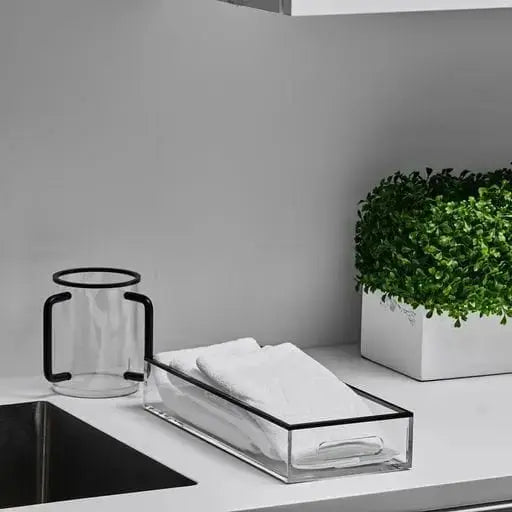 Edge Washing Cup in clear glass with black trim, displayed on white bathroom vanity next to tissues and potted plant