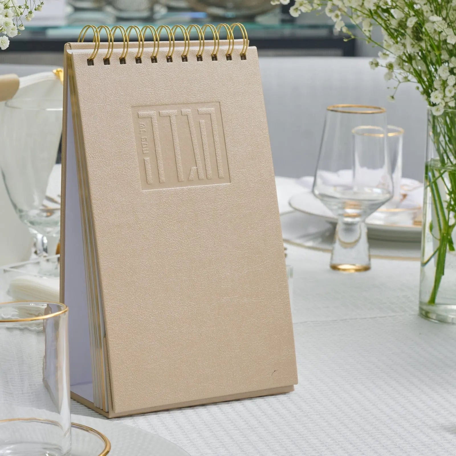 Tan leather standing Haggadah with embossed details displayed on white table with glasses and greenery nearby.