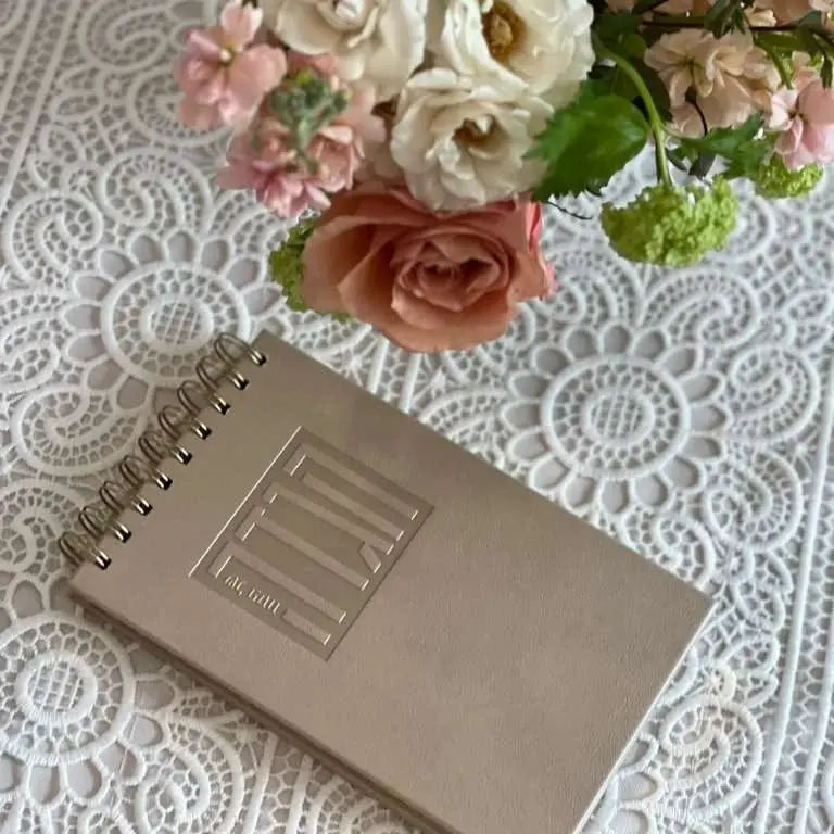 Tan leather painted Haggadah standing on white lace tablecloth with pink roses, Pesach holiday book