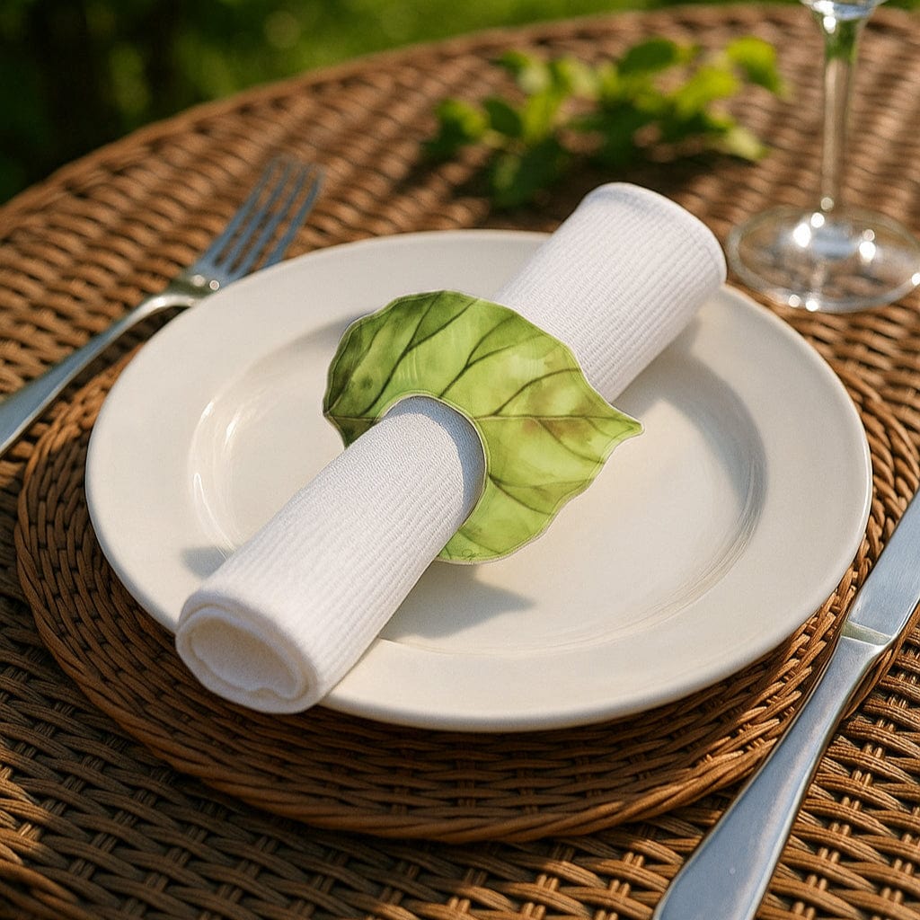 Green Leaf Napkin Rings wrapped around a white linen napkin on an ivory plate, set on a woven rattan outdoor table.