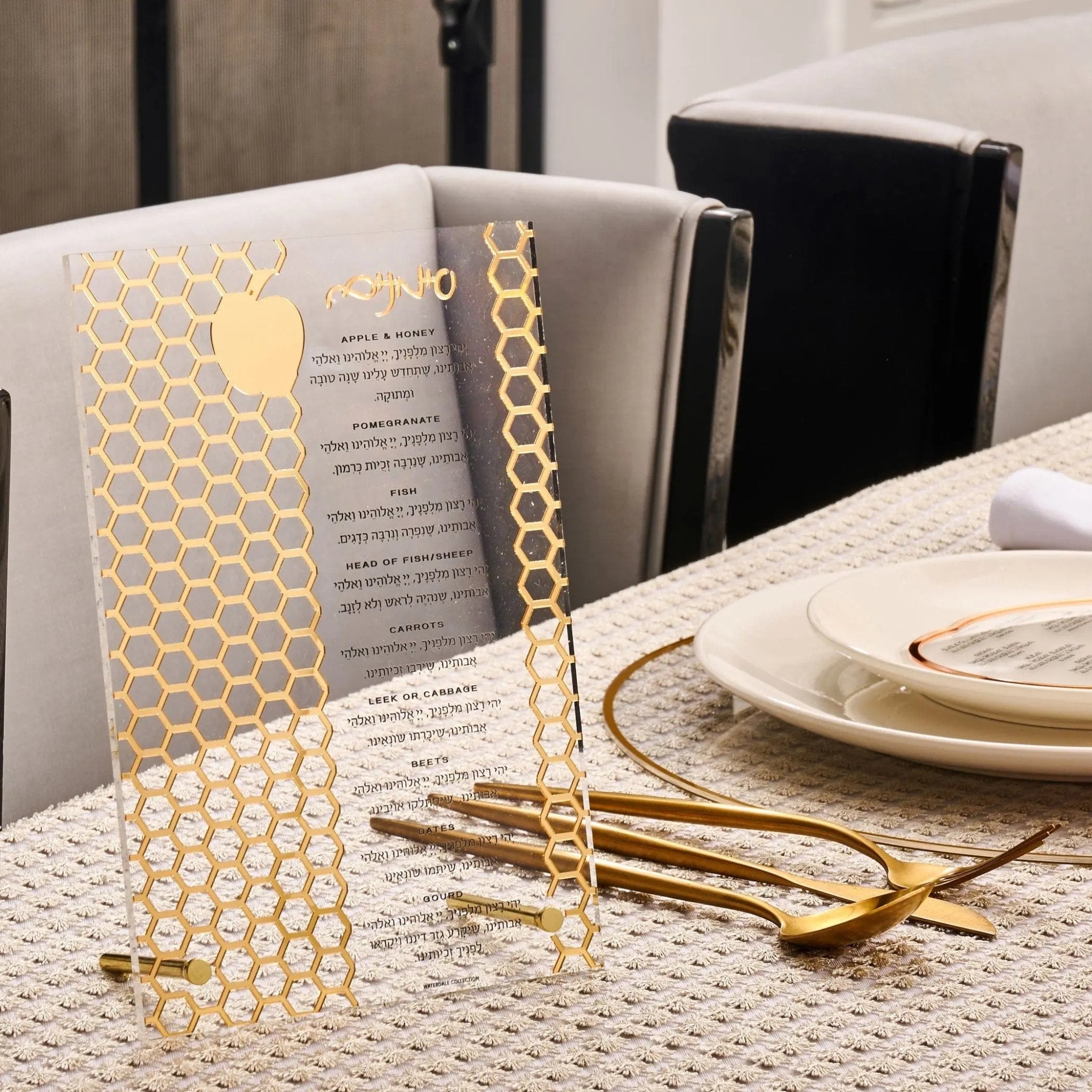 Gold laser cut tabletop Simanim plaque with geometric honeycomb pattern, displayed on white table with place setting and gold utensils.