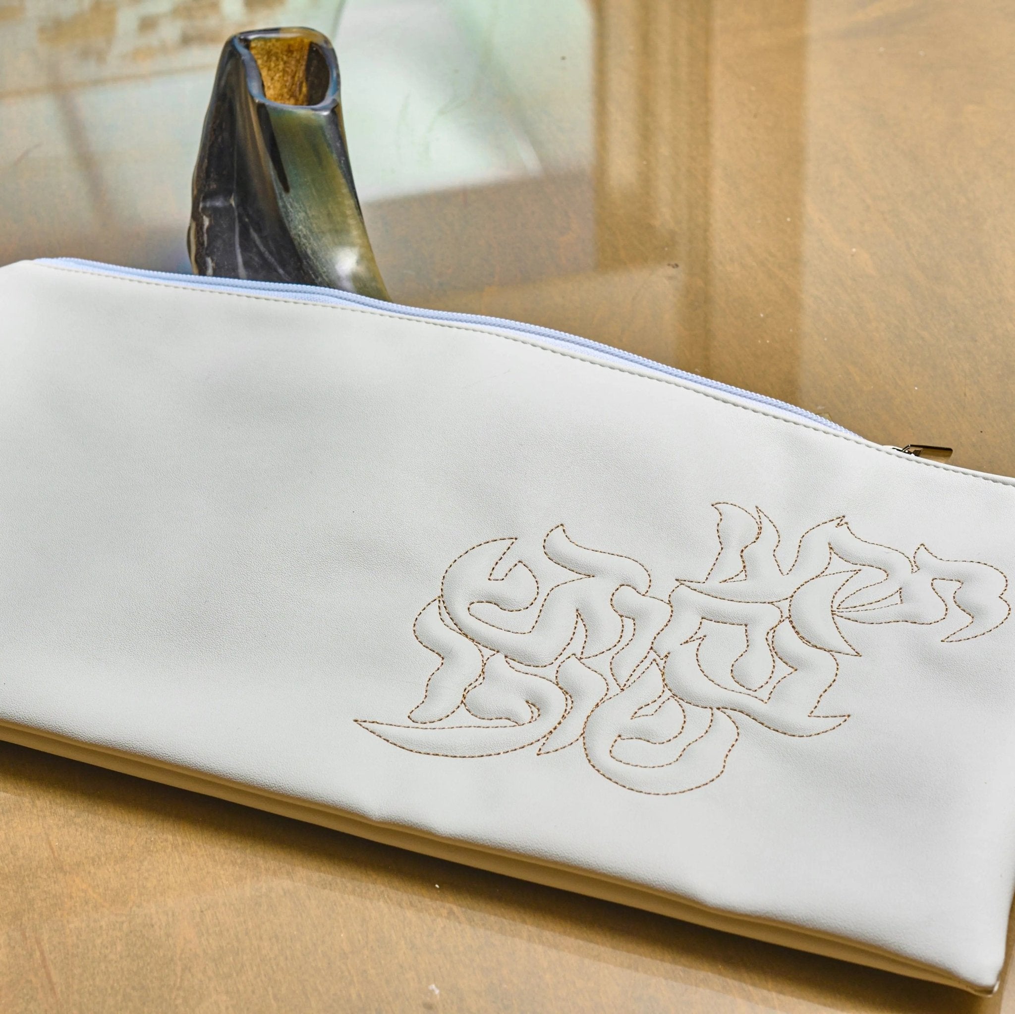 White leather pillow script shofar bag with embossed text design, displayed on wooden surface with gold accent detail visible in background.