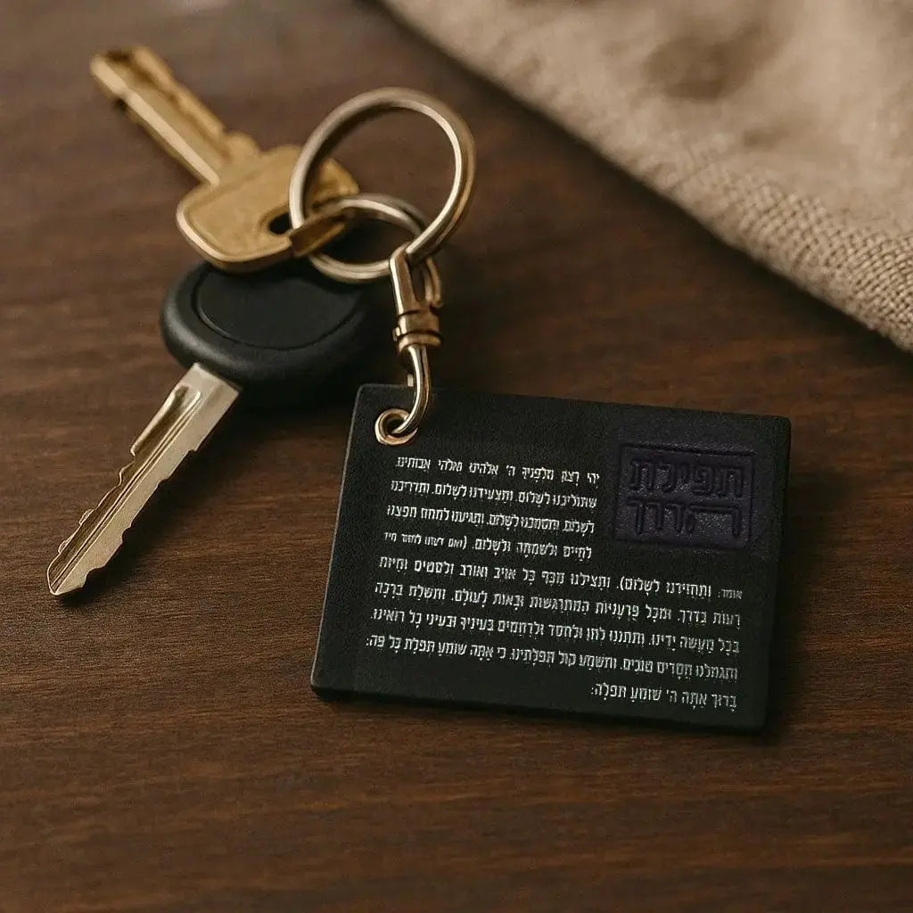 Black leather Tefillas Haderech keychain with gold-tone hardware and Hebrew text card, displayed with two keys on natural linen background.