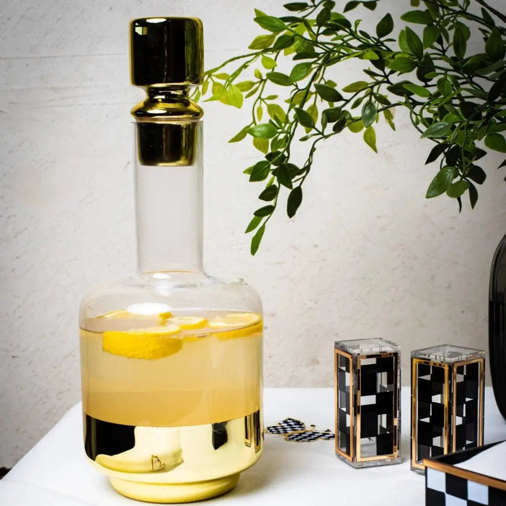 Minimalist wine decanter with clear glass body, yellow wine inside, gold-tone metal stopper and base collar, displayed with green foliage and decorative items.