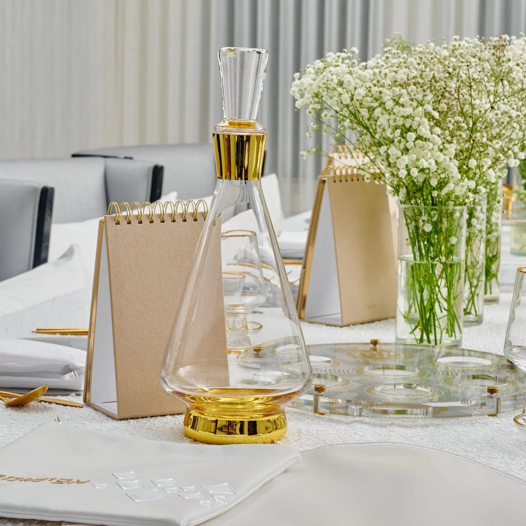 Modern wine decanter in clear glass with gold accents displayed on white marble surface beside fresh flowers and tabletop accessories.