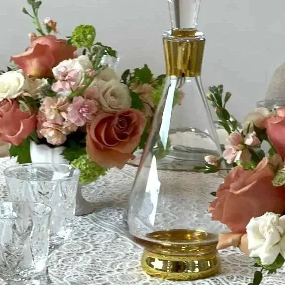 Modern wine decanter with clear glass body and gold stopper, displayed among pink and coral roses in white vases on a marble surface.