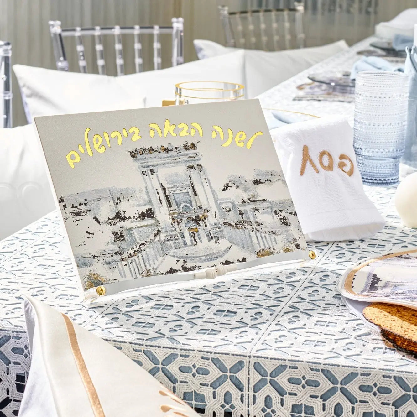 Painted Bais Hamikdash Haggadah stand displaying illustrated Jerusalem design, displayed on table with Passover Haggadah during Pesach seder meal setup.