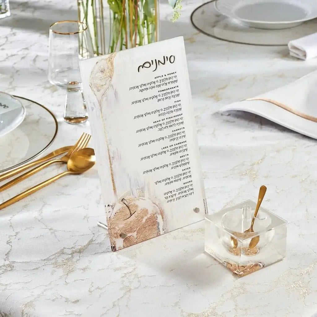 Painted gold tabletop simanim plaques displayed on marble surface with place settings and flowers for Rosh Hashanah holiday table decor.