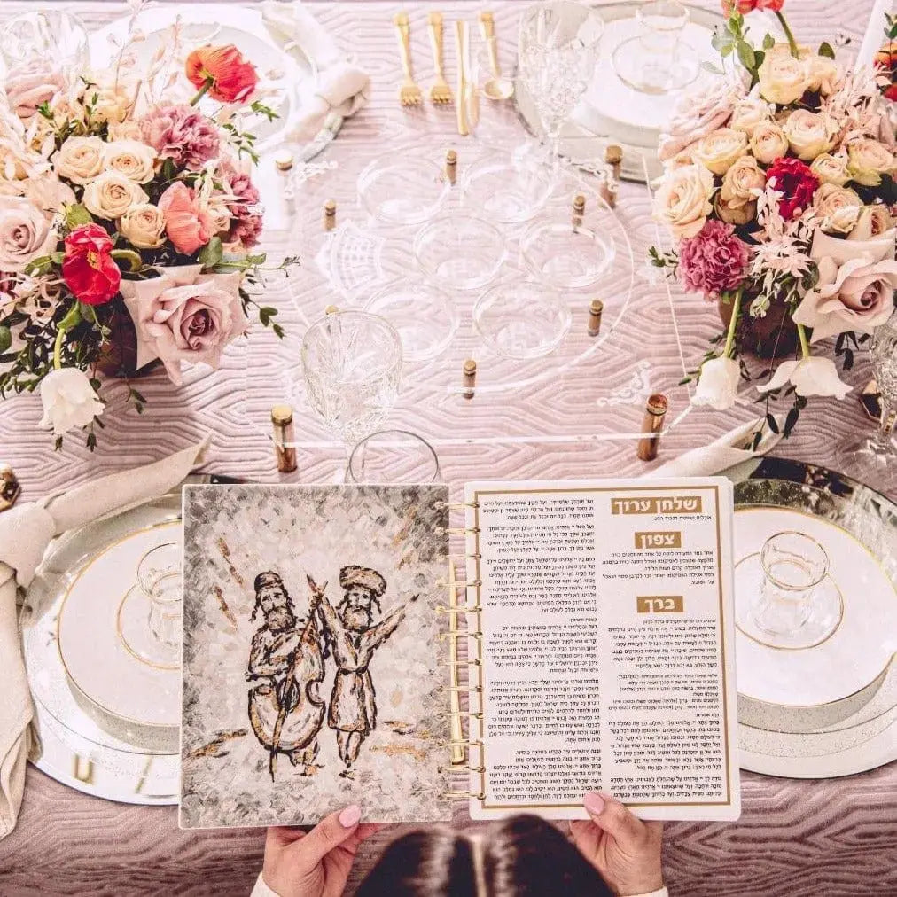 Painted Krias Yam Suf Haggadah displayed on elegant table setting with floral arrangements and place settings for Pesach holiday meal.