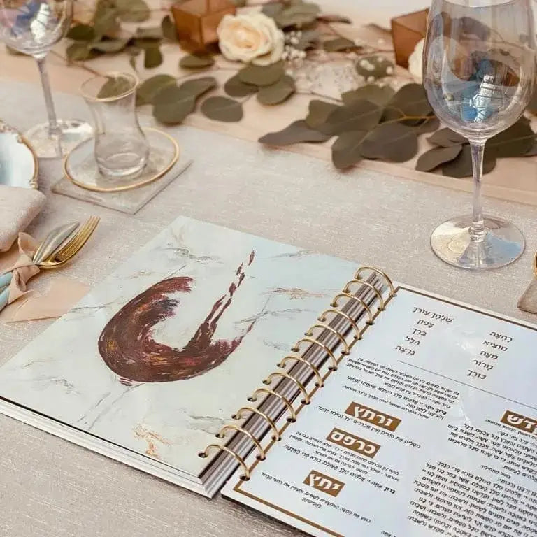 Painted Krias Yam Suf Haggadah displayed on table with eucalyptus, wine glasses, and Pesach planning materials
