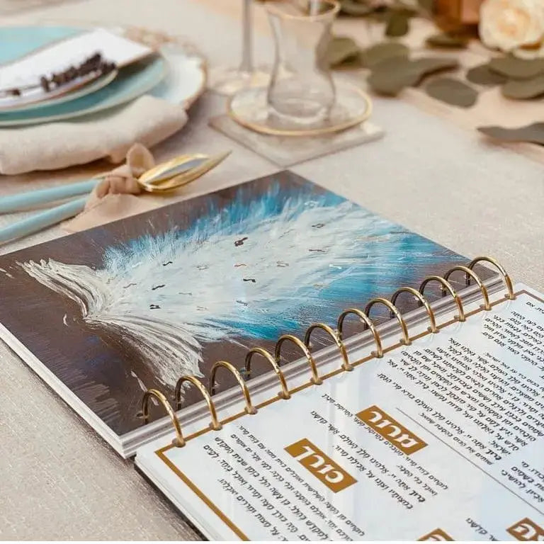 Painted Krias Yam Suf Haggadah with blue watercolor cover art displayed on table with place settings and Pesach materials