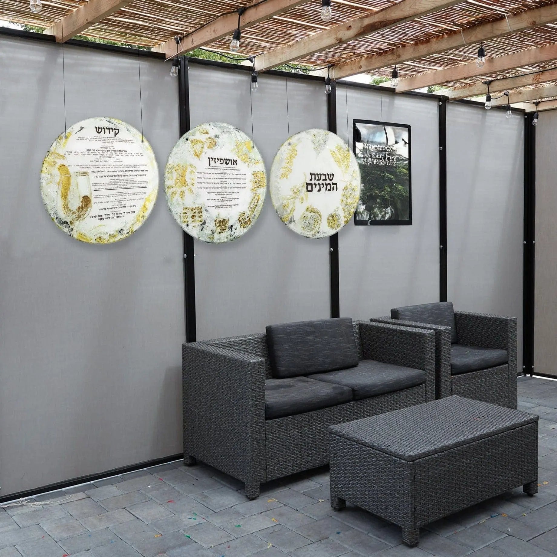 Painted Sukkah decoration set featuring three round pendant lanterns with botanical designs hanging in a modern sukkah booth with gray furniture.