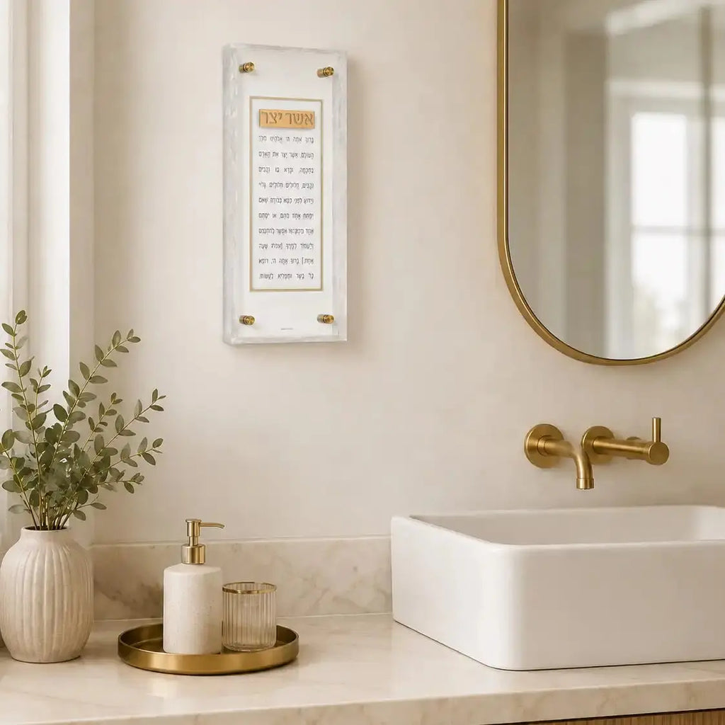 Pearlstone Frame Asher Yatzar wall art displayed above a white bathroom sink with brass fixtures and marble countertop.