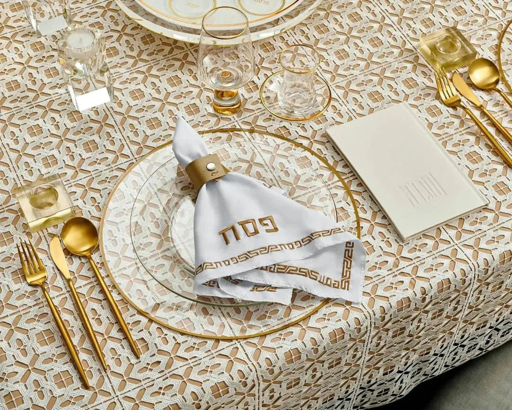 Gold leather Pesach napkin rings arranged on a beige textured tablecloth with gold cutlery and place settings for holiday dining.