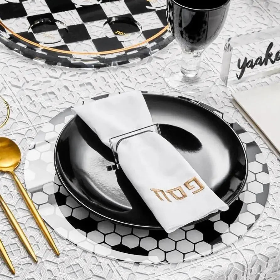 Black and white checkered Pesach napkin ring displayed on a folded white napkin with gold utensils and tableware accents.